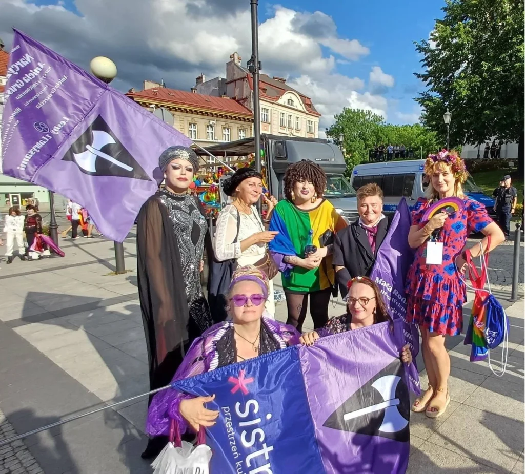 Group of lesbians from Sistrum in Poland, some in drag posing with purple lesbian flag, smiling for a photo